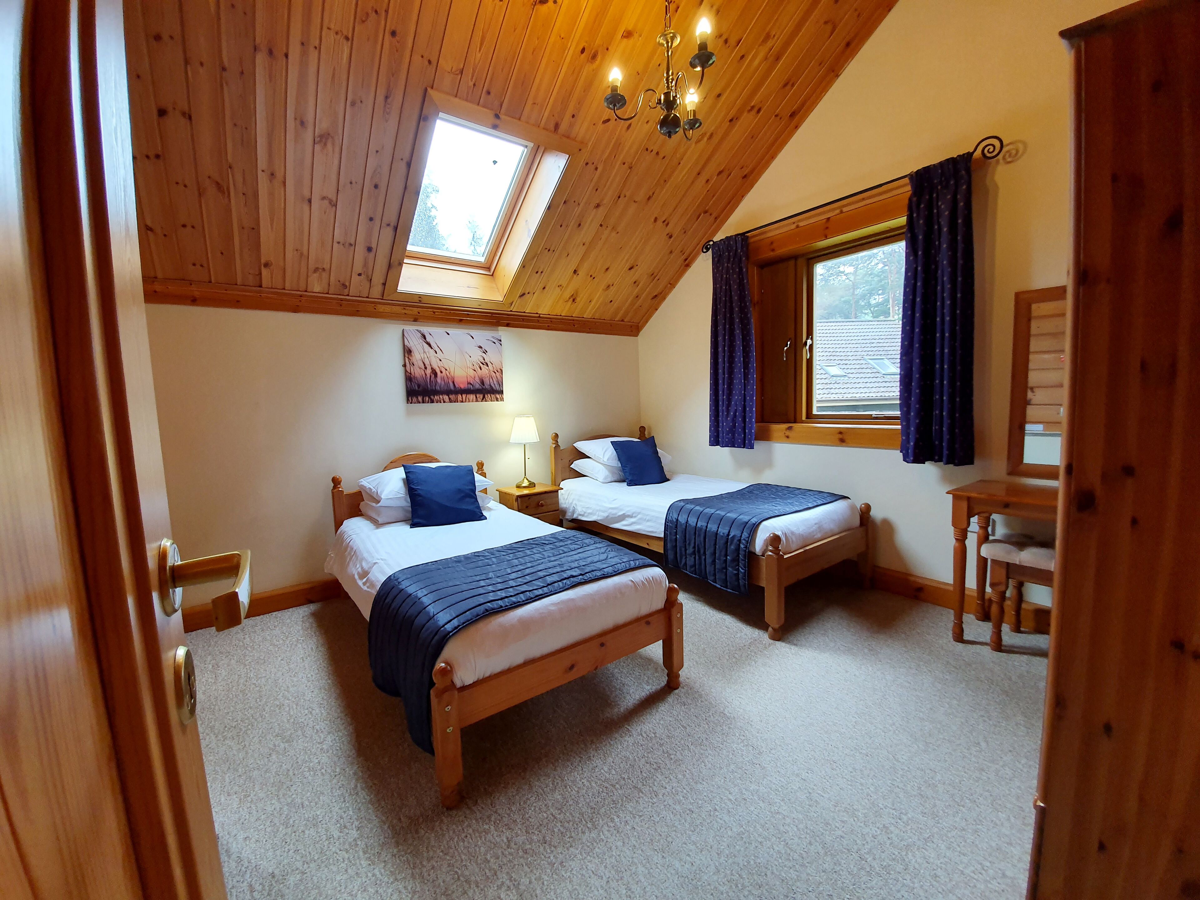 A cozy room with two wooden beds, a desk, a chair, a lamp, and a painting on the wall.