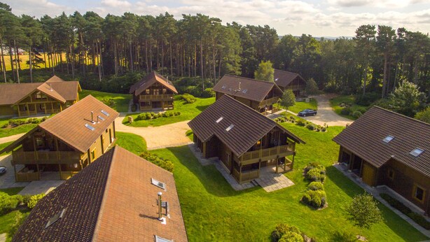 A cluster of wooden cabins with sloped roofs, surrounded by well-maintained lawns and a forested backdrop. - The Dorset Resort (Wareham)