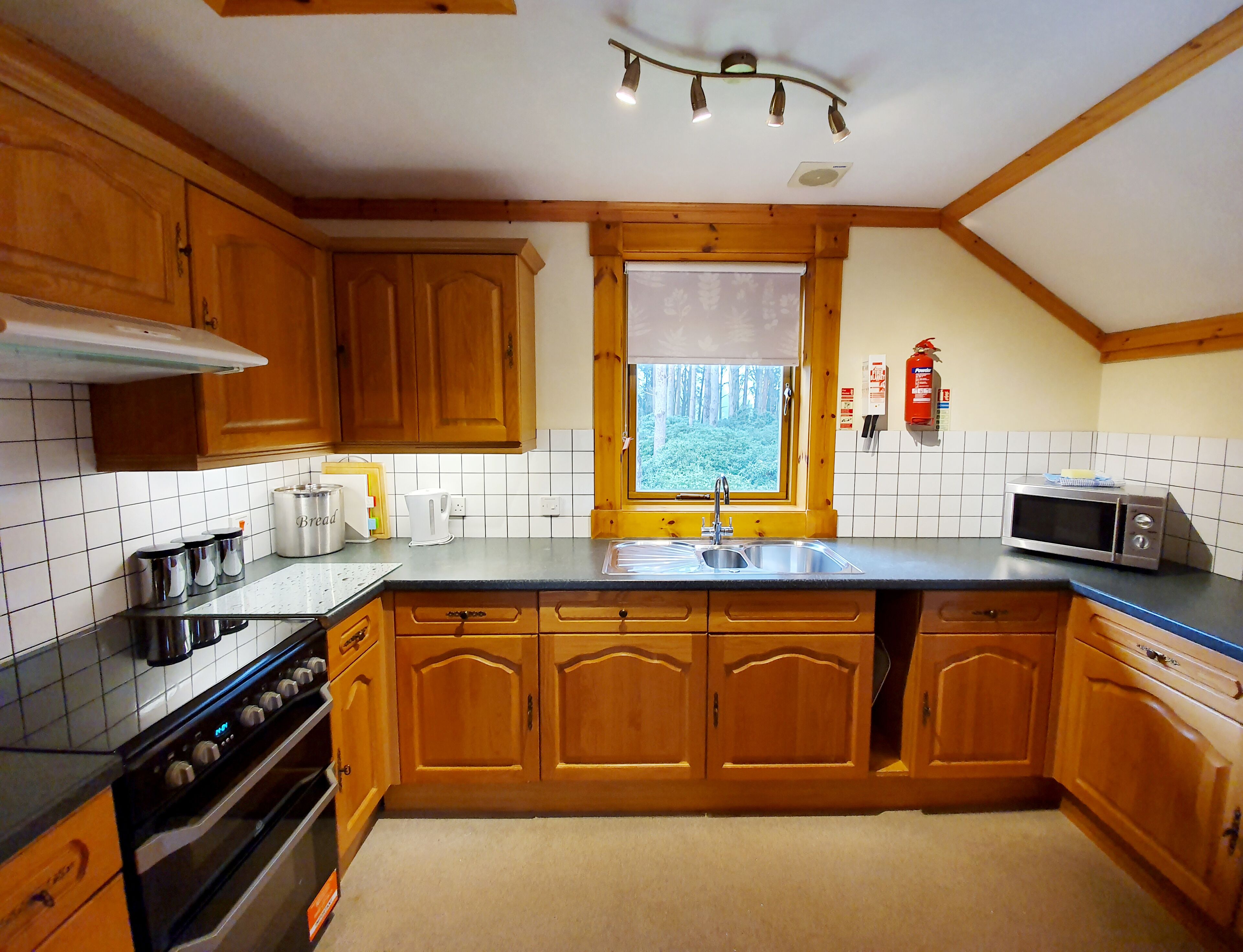 A kitchen with wooden cabinets, a stainless steel sink, a microwave, and a window with a view of trees.