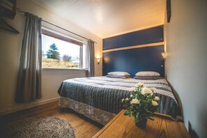 A bedroom with a bed, a window with curtains, a bedside table with a vase of flowers, and a view of the outdoors.
