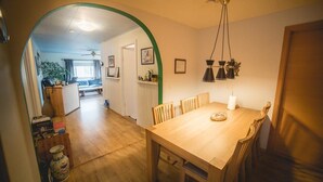 Een eetruimte met een houten tafel en stoelen, een hanglamp en uitzicht op een andere kamer met een plafondventilator.