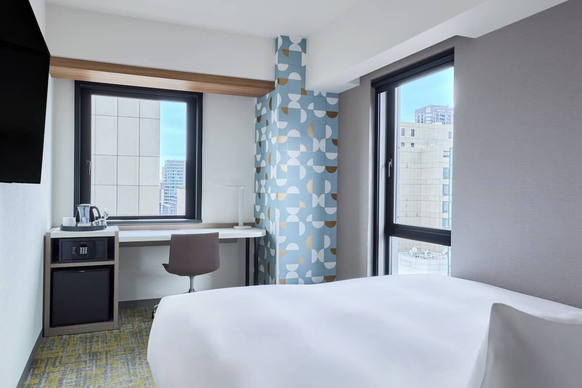 A hotel room with a bed, a desk with a chair, a microwave, and a window with a city view.
