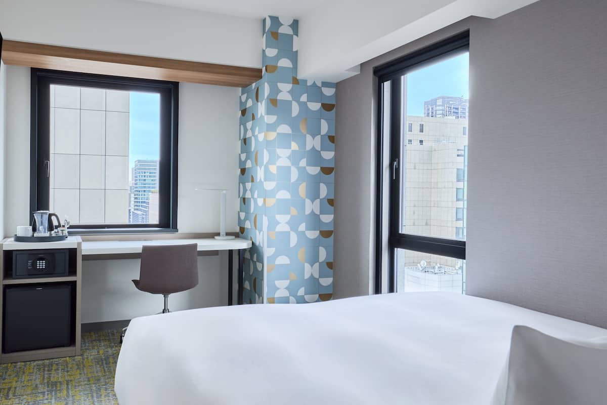 A hotel room with a bed, desk, chair, and a view of the cityscape through large windows.