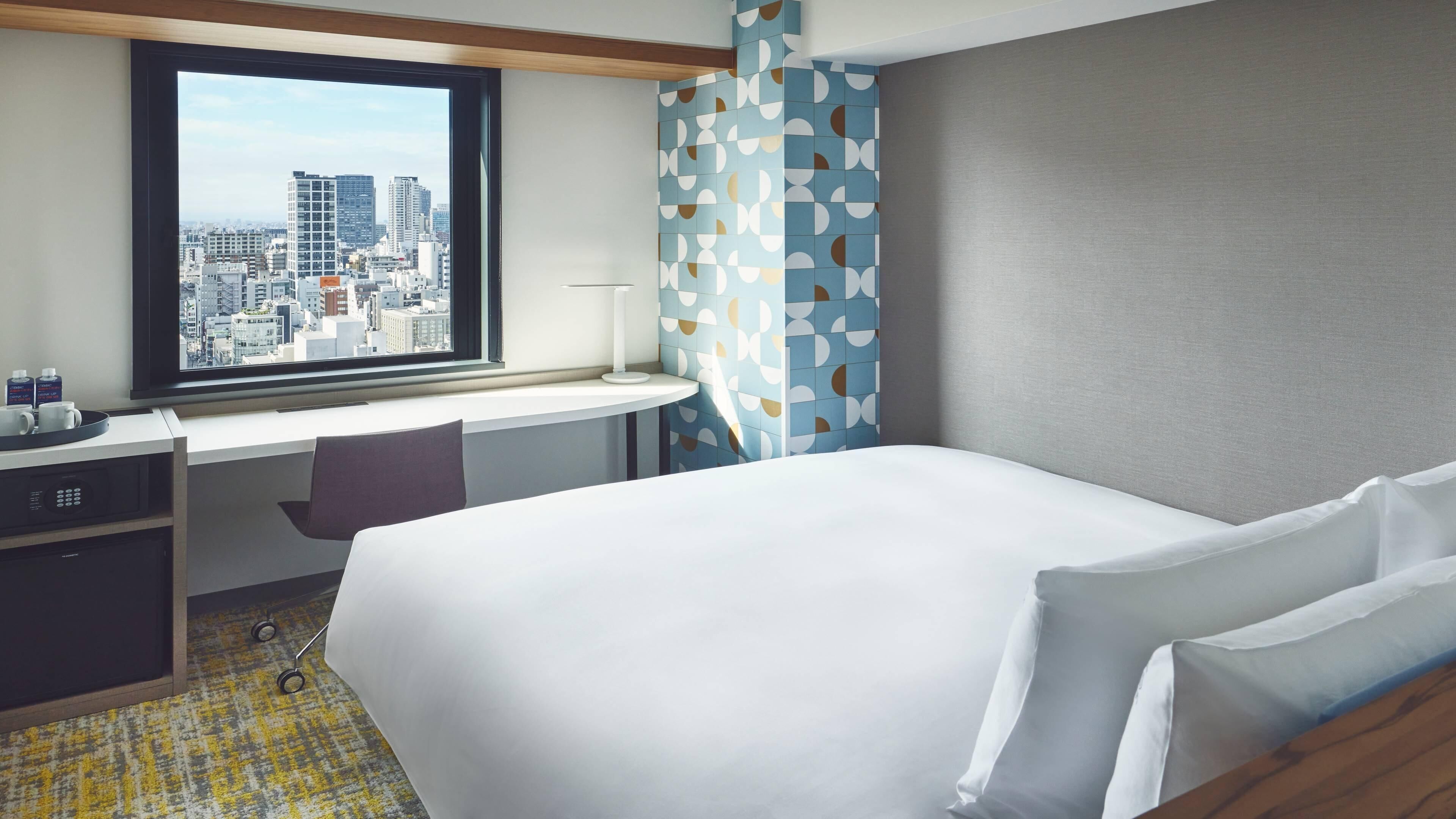 A hotel room with a bed, a desk with a chair, a window with a city view, and a patterned curtain.