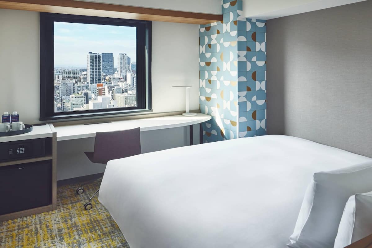 A hotel room with a bed, a desk with a chair, a window with a city view, and a patterned curtain.