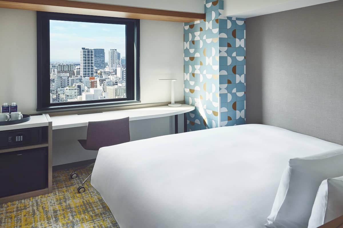 A hotel room with a bed, a desk with a chair, a window with a city view, and a patterned curtain.