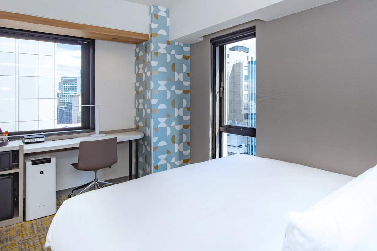 A hotel room with a bed, desk, chair, and a large window offering a city view.