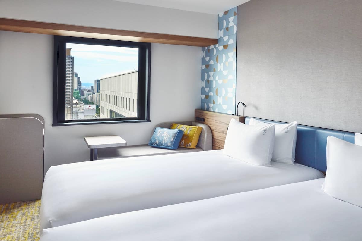 Habitación de hotel con una cama grande, una mesita de noche pequeña, una silla y una ventana con vistas a la ciudad.