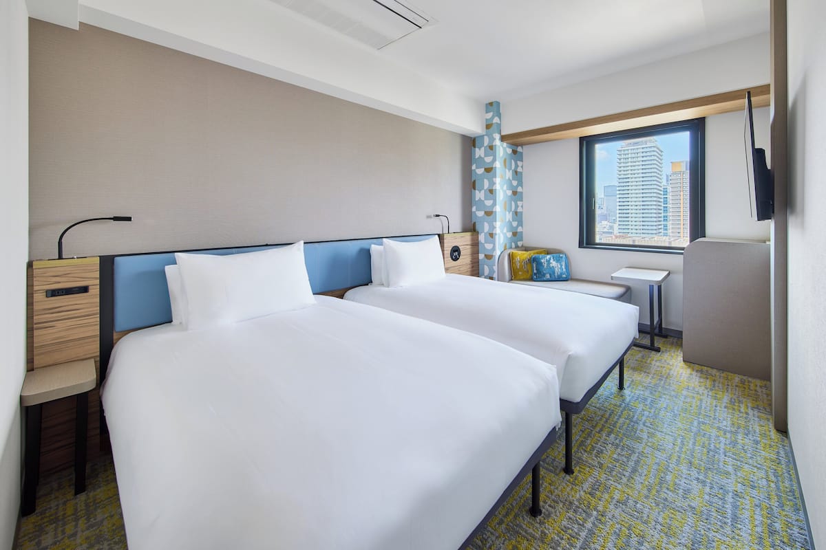 A hotel room with two beds, a small table, a chair, a TV, and a window with a city view.