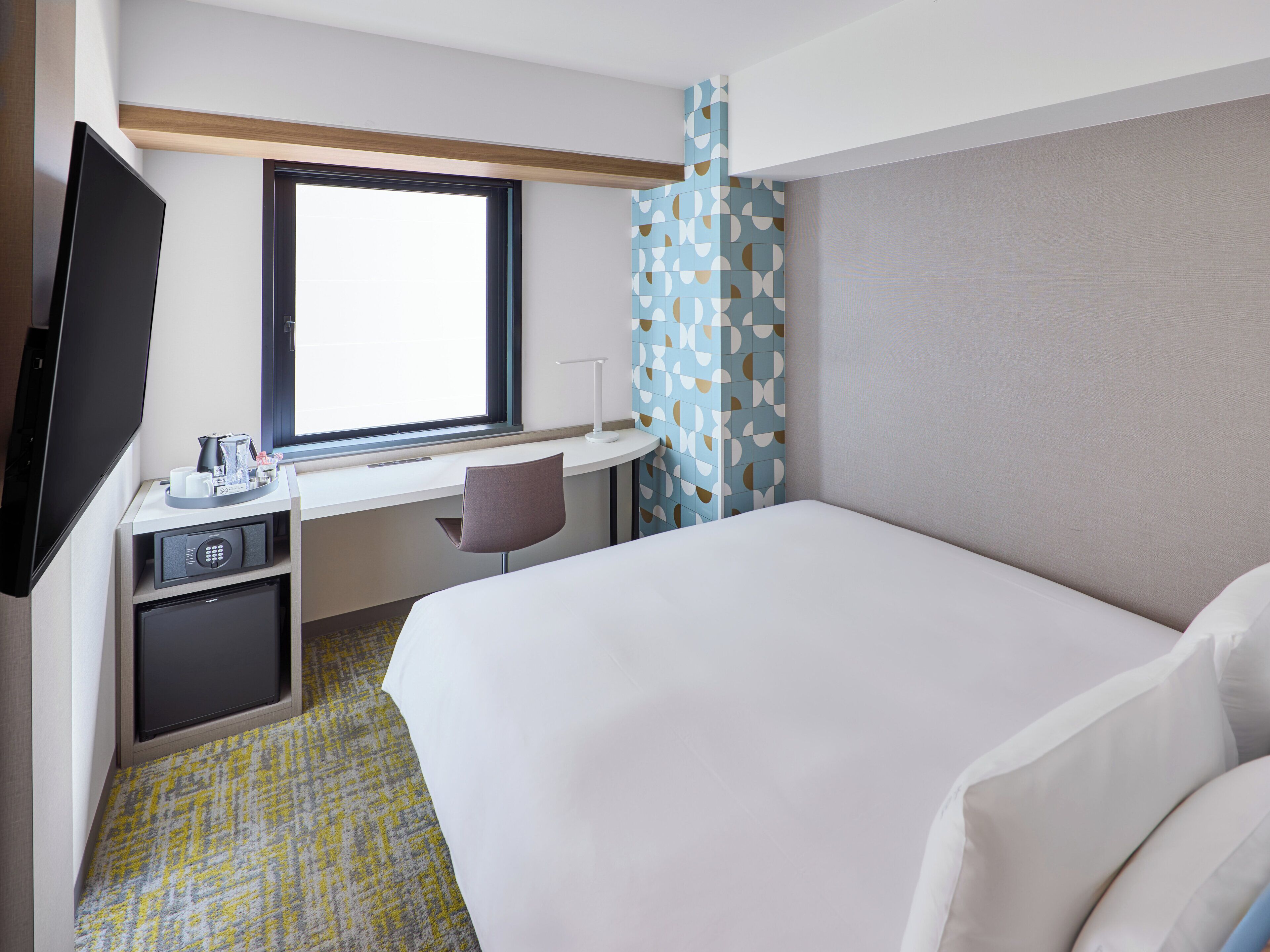 Quarto de hotel com cama, escrivaninha, cadeira, televisão e janela.
