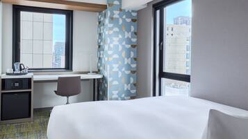 A hotel room with a bed, desk, chair, and a view of the cityscape through large windows.
