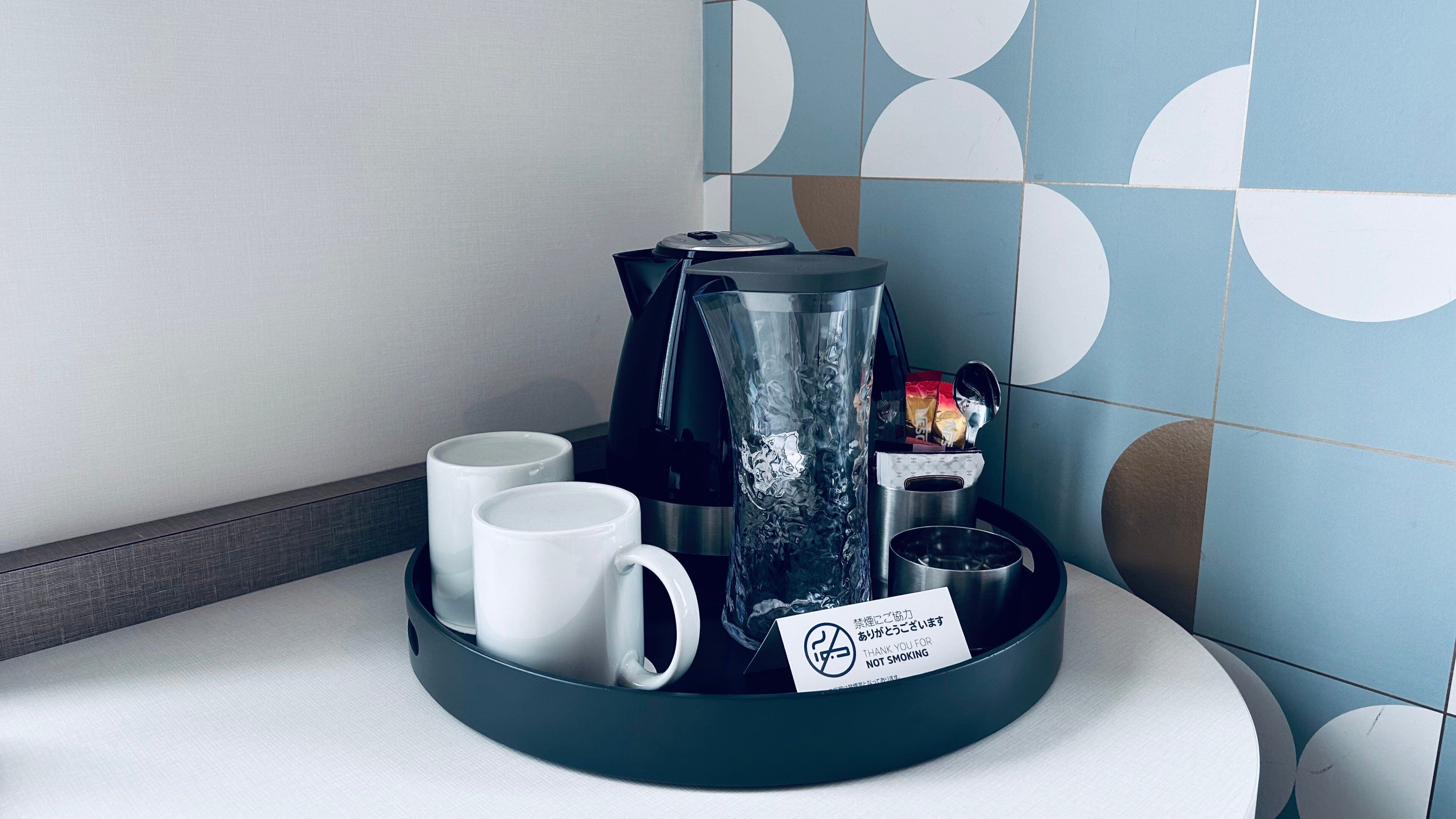 A coffee station with a coffee maker, two white mugs, and a tray with a 'No Smoking' sign.