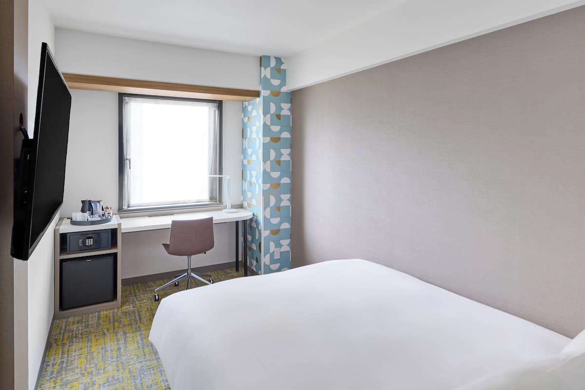 A hotel room with a bed, a desk with a chair, a television, and a window.