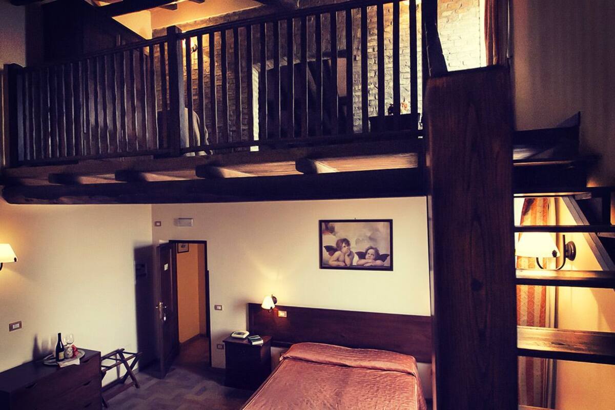 Ein Hotelzimmer mit erhöhter Schlafkoje, einem Bett mit roter Decke, einem Nachttisch mit Lampe, einer Holzkommode und einem Bild an der Wand.