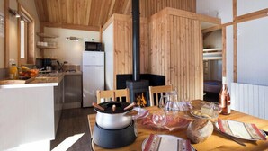 Un interior acogedor de cabaña, con revestimiento de madera, una chimenea central, una mesa de comedor dispuesta con comida y bebidas, y una zona de cocina con un refrigerador y estanterías.