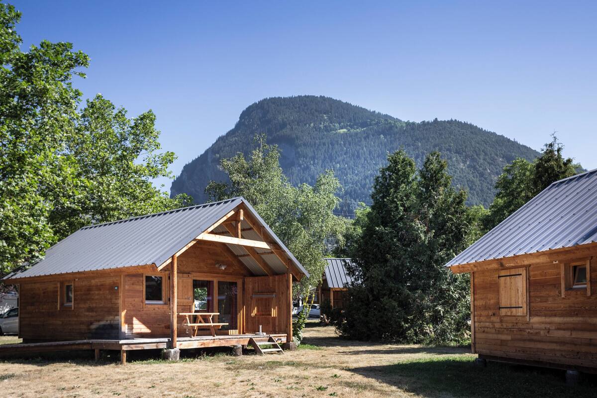 A wooden cabin with a porch and a dining table, surrounded by trees and a mountain in the background.