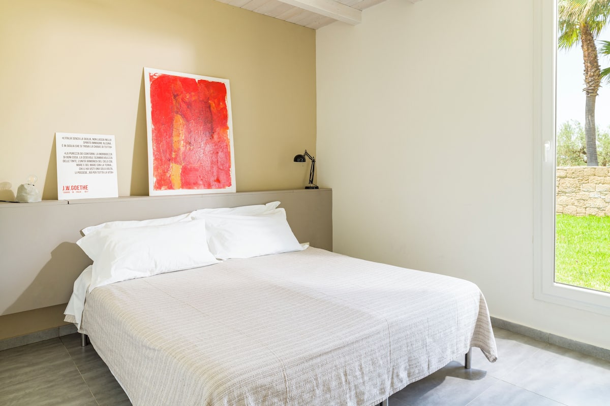 A bedroom with a bed, a red abstract painting, a desk with a lamp, and a window with a view of greenery.