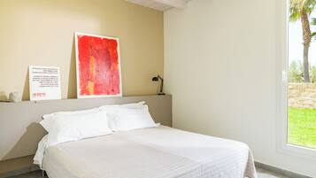 A bedroom with a bed, a red abstract painting, a desk with a lamp, and a window with a view of greenery.