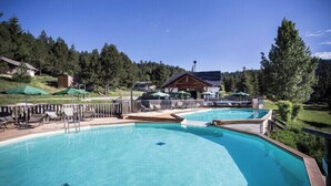 A swimming pool with lounge chairs, a wooden deck, and a building in the background. - Huttopia Font Romeu (Font-Romeu-Odeillo-Via)