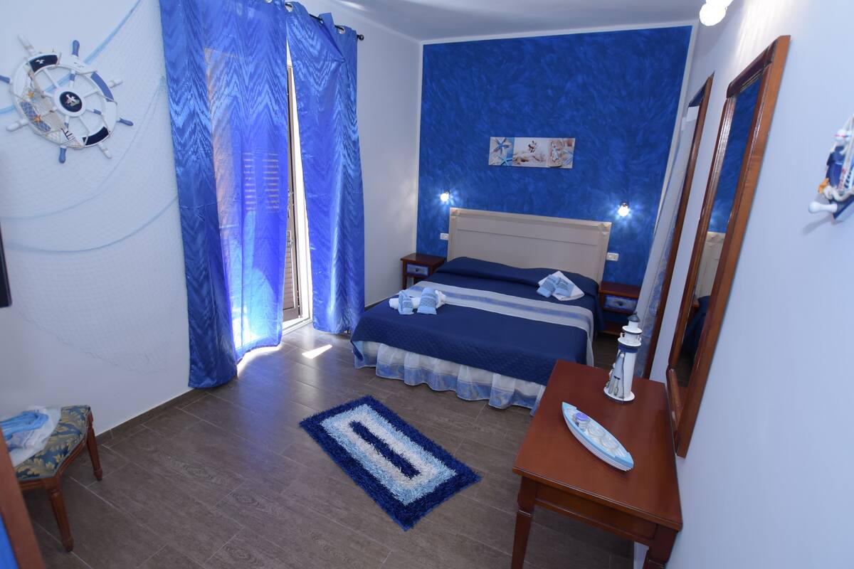 A bedroom with a bed, a nightstand, a wooden dresser, a desk, a chair, a blue and white rug, a window with blue curtains, a wall clock, and a wall-mounted mirror.