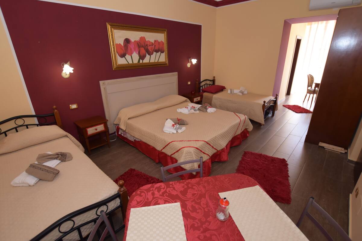 A hotel room with two beds, a dining table, and a chair. There is a painting on the wall and a red rug on the floor.