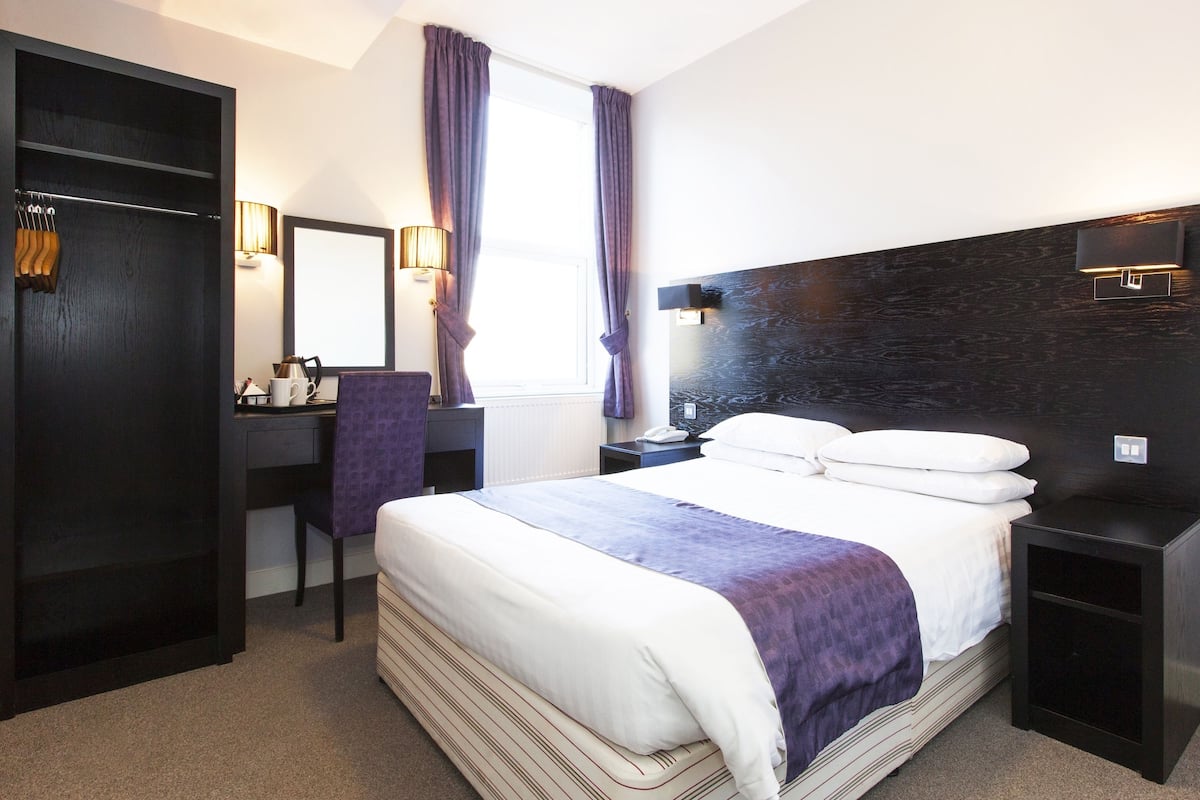 A hotel room with a large bed, a desk with a chair, a wardrobe, and a nightstand with a lamp.