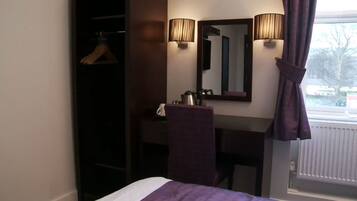 A hotel room with a bed, a desk, a mirror, a hanging coat rack, and a window with curtains.