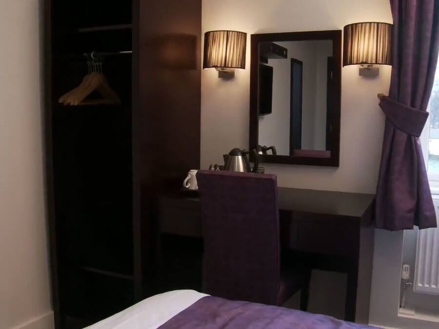 A hotel room with a bed, a desk, a mirror, a hanging coat rack, and a window with curtains.