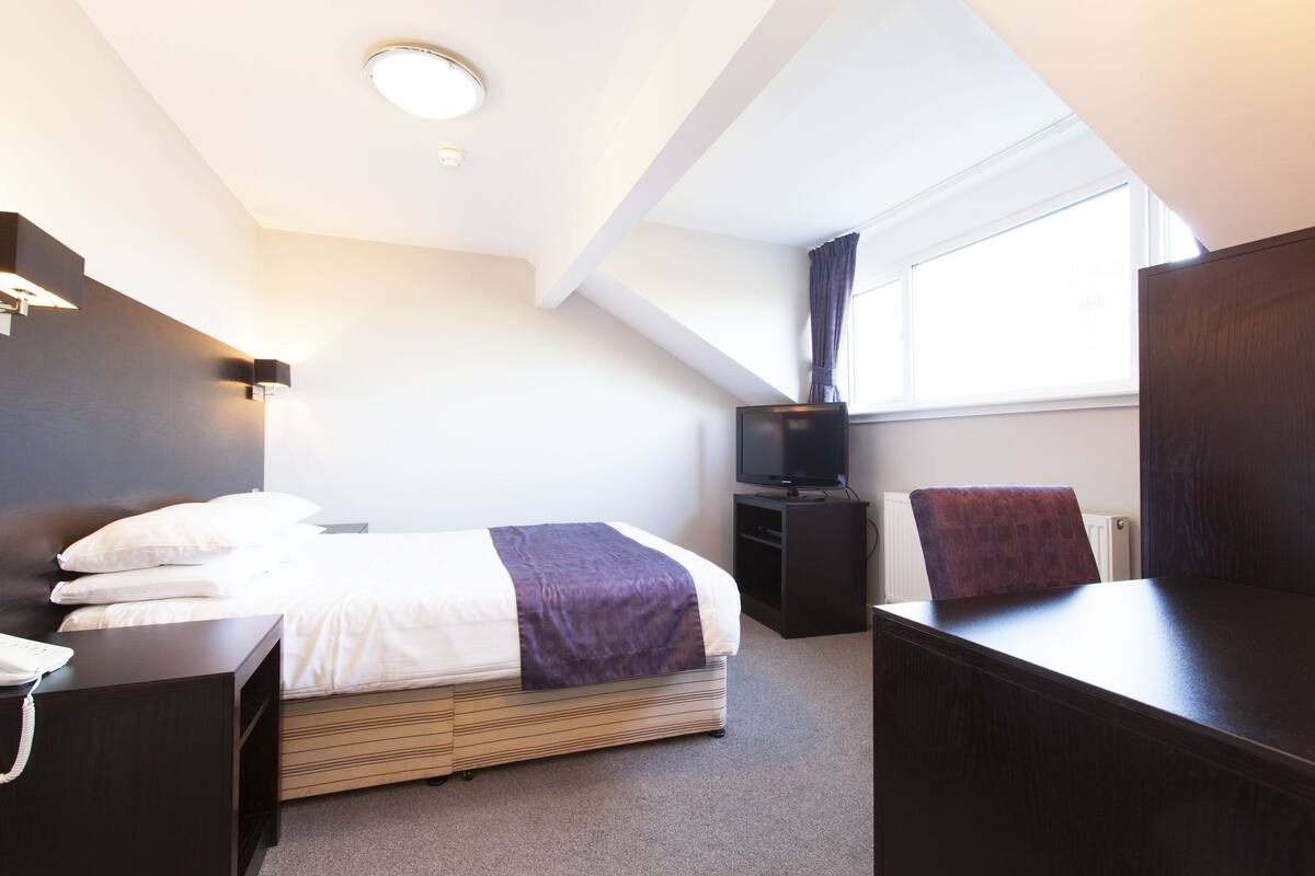 A hotel room with a bed, bedside table, TV, and a desk with a chair.