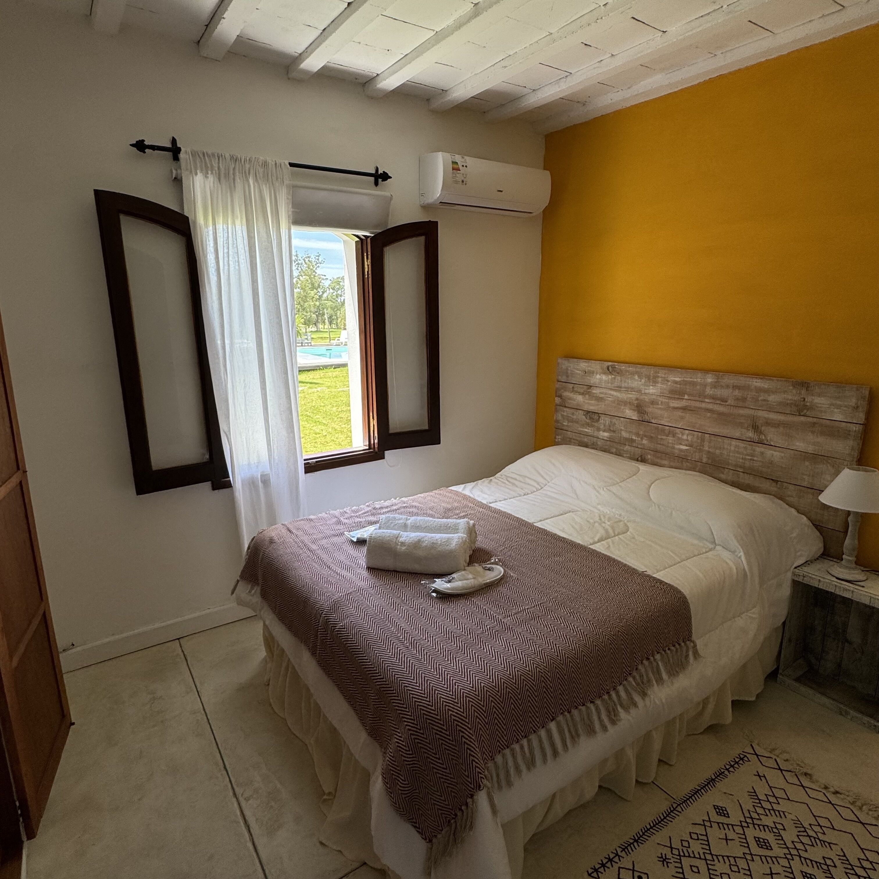 A bedroom with a bed, a window with curtains, an air conditioning unit, and a bedside table with a lamp.