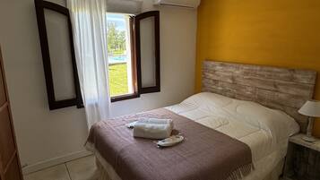 A bedroom with a bed, a window with curtains, an air conditioning unit, and a bedside table with a lamp.
