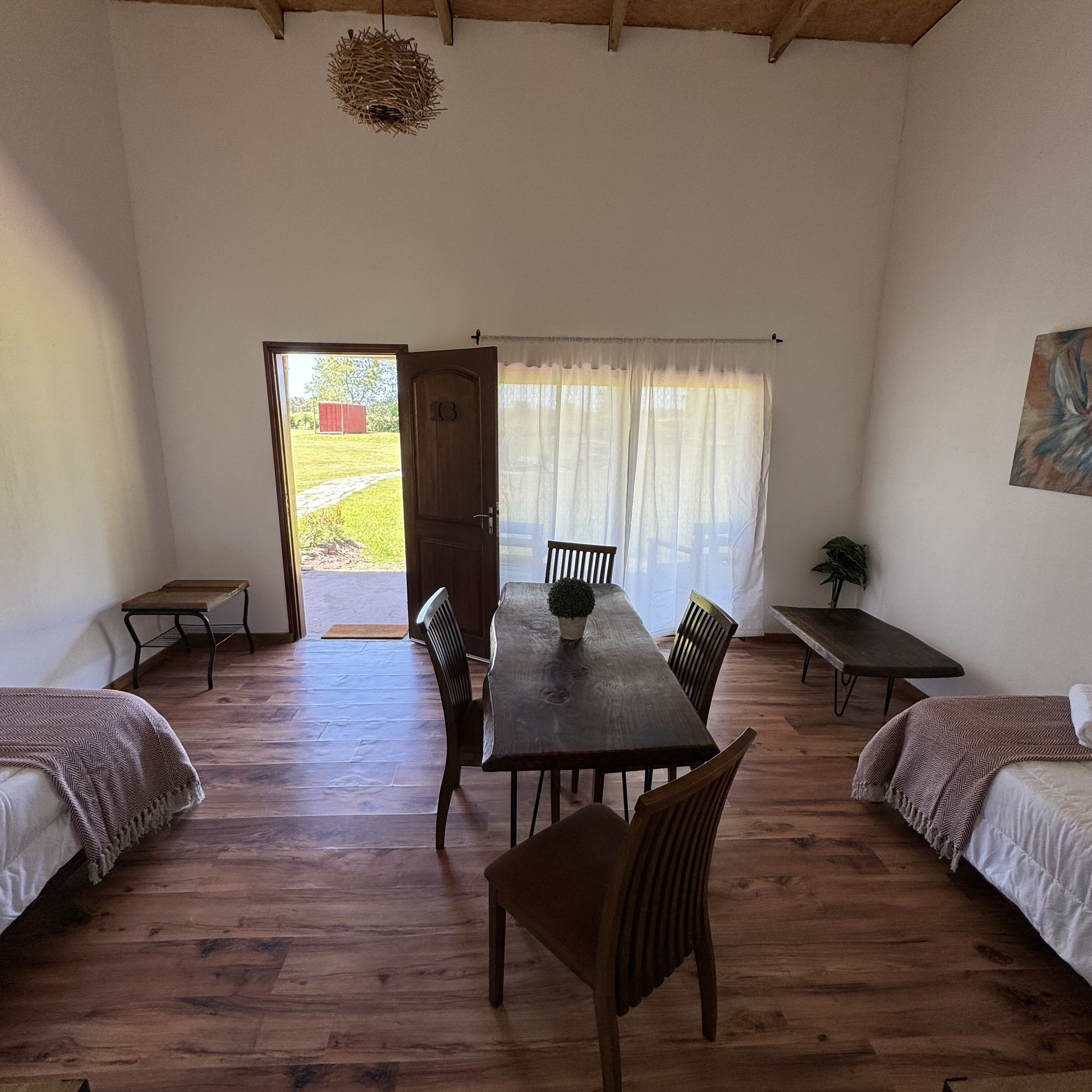 A room with a wooden floor, a dining table with chairs, two beds with blankets, a door leading outside, and a hanging light fixture.