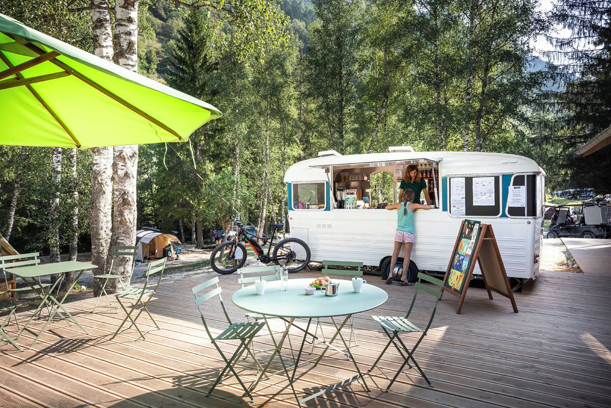 Un camion-restaurant mobile se trouve dans une zone boisée, avec une table et des chaises, un vélo et une tente en arrière-plan.