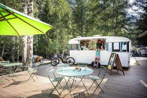 A mobile food truck in a forested area with a table and chairs, a bicycle, and a tent in the background.