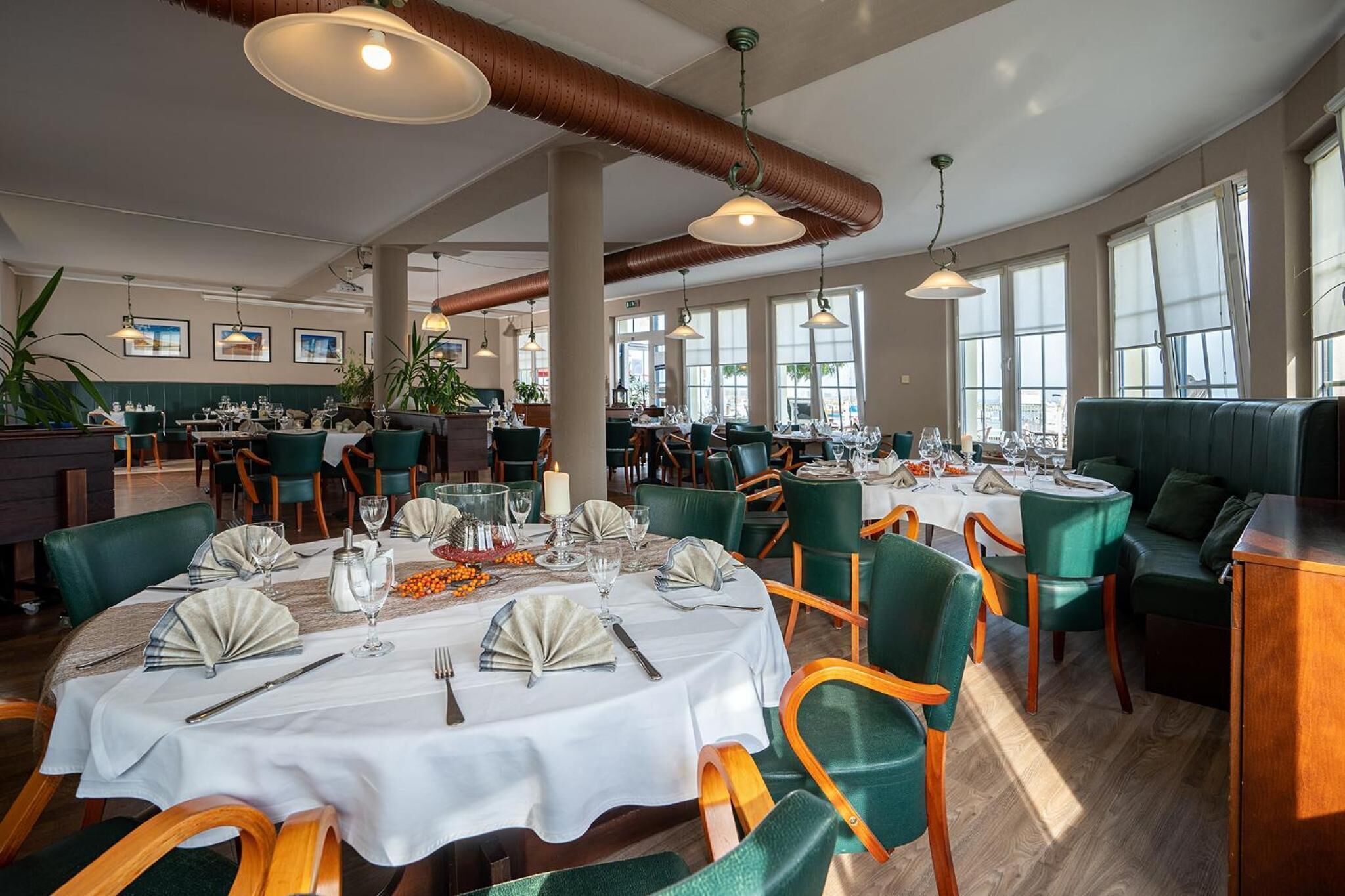 A restaurant with round tables set for dining, green upholstered chairs, and large windows allowing natural light.
