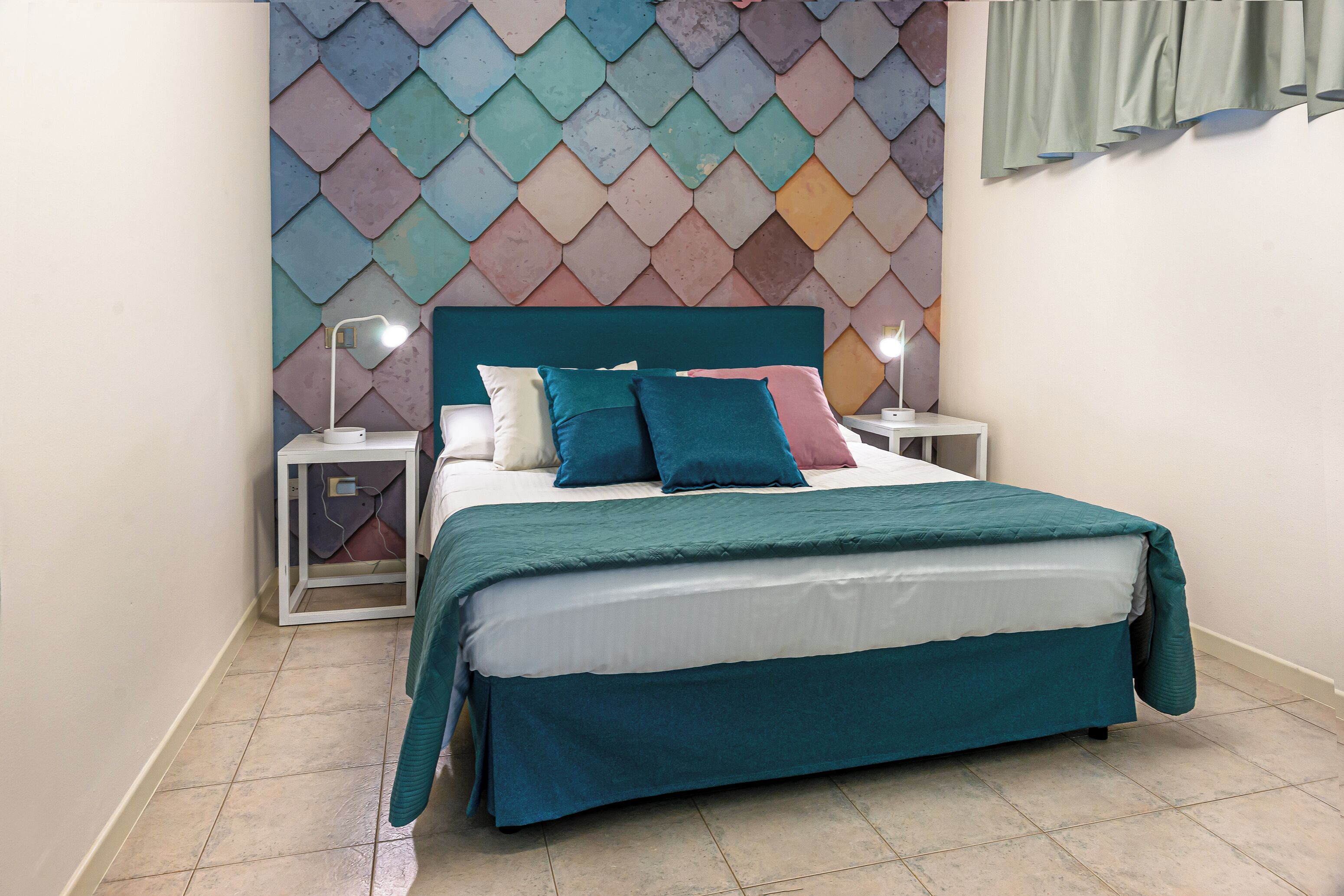 Ein ordentlich bezogenes Bett mit türkisfarbenem Bettbezug und Kissen, zwei Nachttische mit Lampen sowie eine farbenfrohe Wand mit geometrischem Muster.