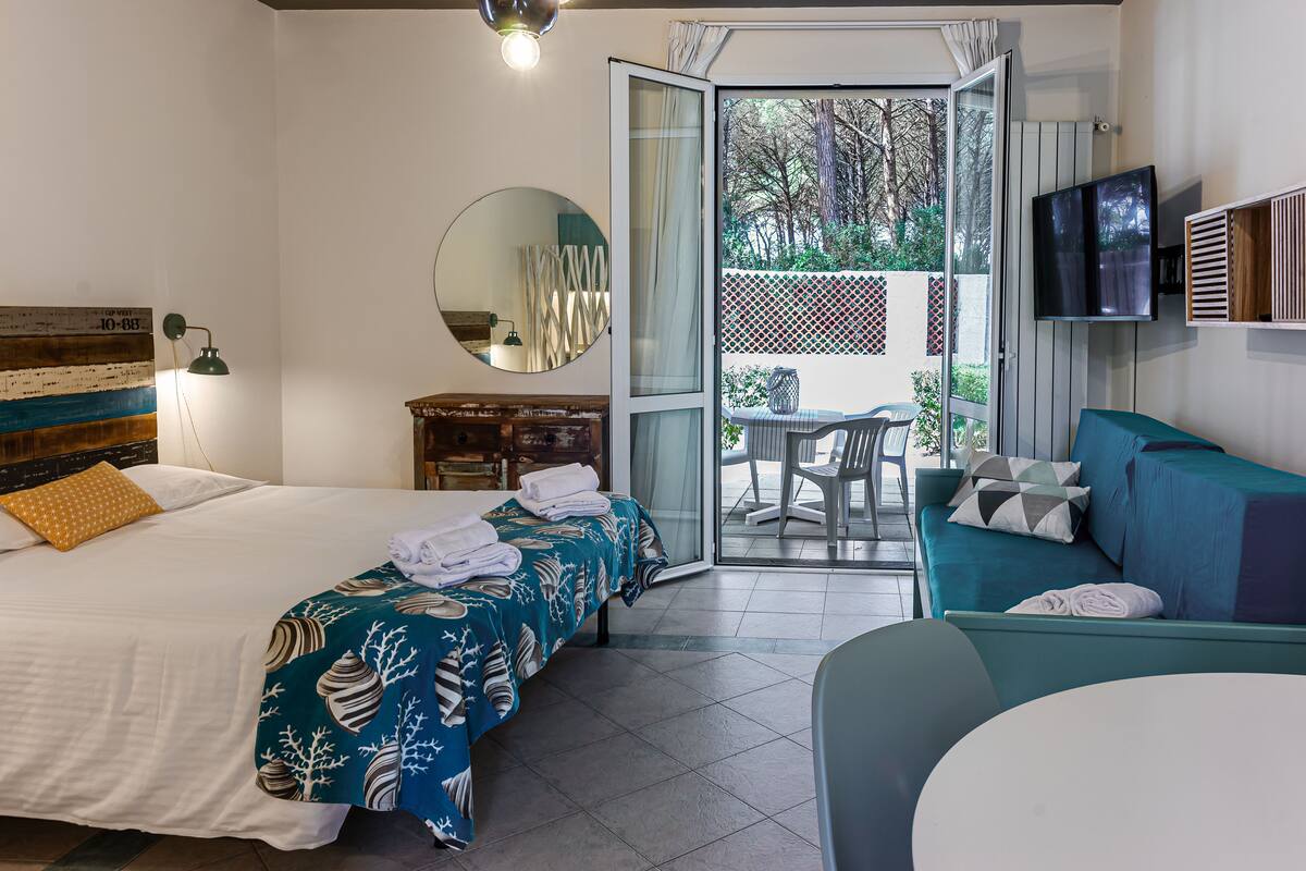 A hotel room with a bed, a sofa, a TV, and a balcony with a view of trees.