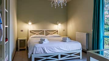Un dormitorio con una cama blanca, dos mesitas de noche, una araña, una ventana con cortinas y un radiador.