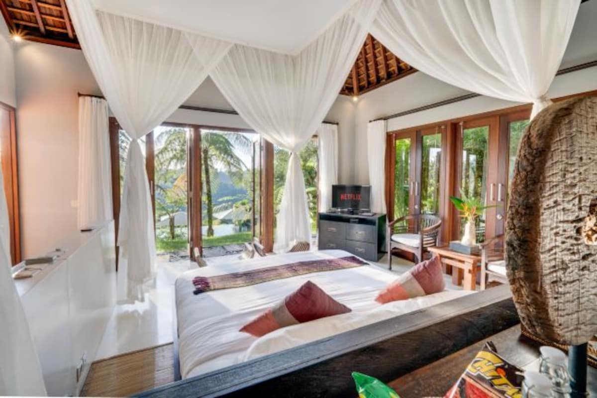 A bedroom with a bed, curtains, a TV, and a view of greenery outside.