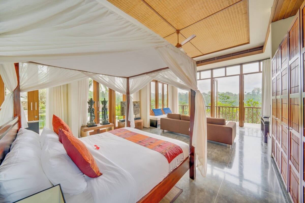 A spacious bedroom with a canopy bed, a sitting area with a sofa and chairs, and large windows offering a view of the outdoors.