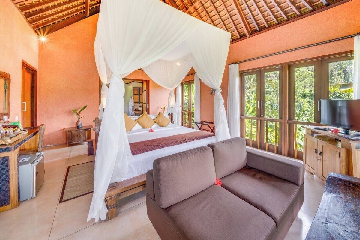 A bedroom with a canopy bed, a sofa, a TV, and a view of greenery.