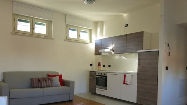 A modern kitchen with white cabinets and a built-in oven. There is a grey sofa with red cushions in the living area. - Appartamento Renzo (Lecco)