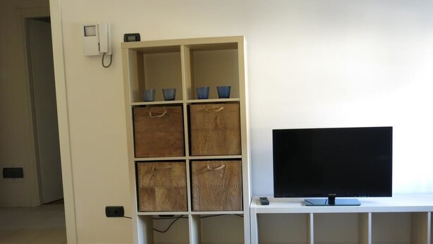 A living room with a TV on a stand, a storage unit with baskets, and a striped rug. - Appartamento Don Abbondio (Lecco)