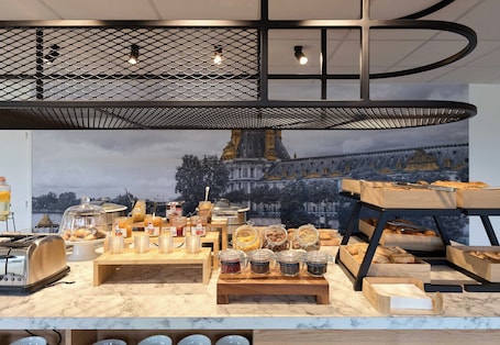 A breakfast buffet with various food items and condiments, a toaster, and a large window with a view of a building.. B&B HOTEL Paris Vitry-sur-Seine 3 étoiles