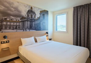 Hotellihuone, jossa on suuri sänky, sängynpääty, jossa on kaupunkimaisemaa kuvaava seinämaalaus, ikkuna verhoilla ja yöpöytä lampulla.