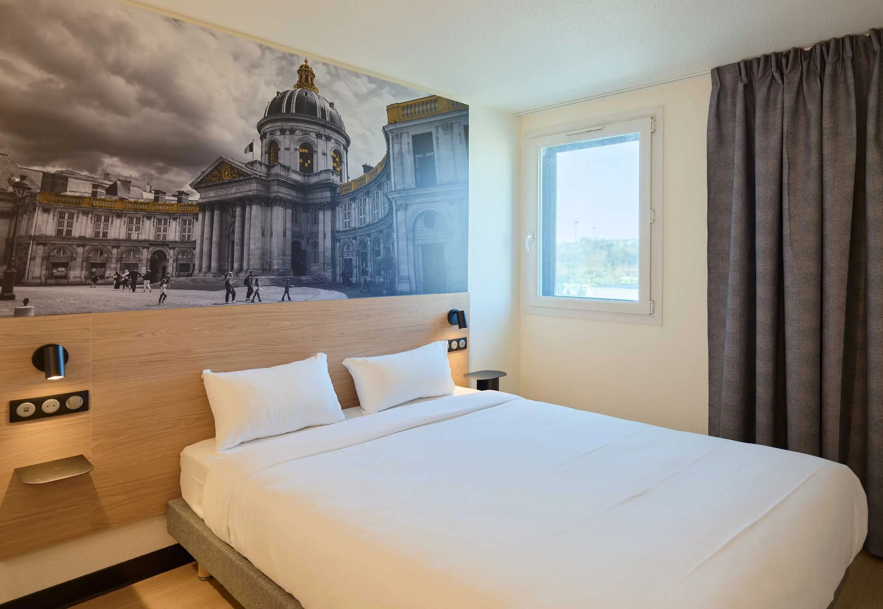 A hotel room with a large bed, a headboard with a mural, a bedside table, a nightstand, a window with curtains, and a view of the sky.