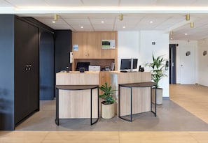 A reception area with a wooden counter, two round tables, a potted plant, and a dark cabinet. - B&B HOTEL Paris Vitry-sur-Seine 3 étoiles (Vitry-sur-Seine)