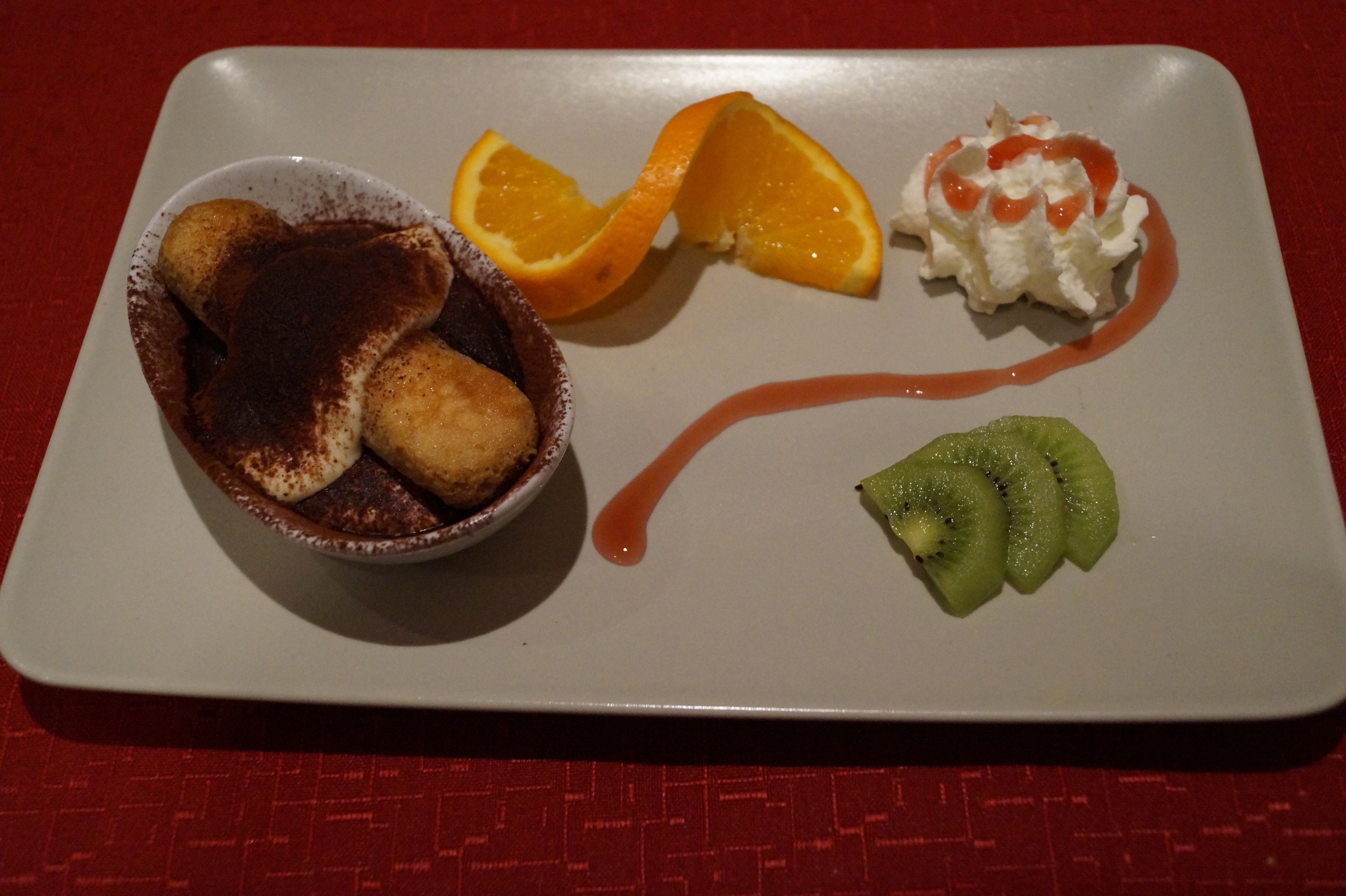 Un plato de postre con un tazón que parece contener un postre de chocolate, una rodaja de naranja, una cucharada de crema batida con salsa, y kiwi en rodajas.