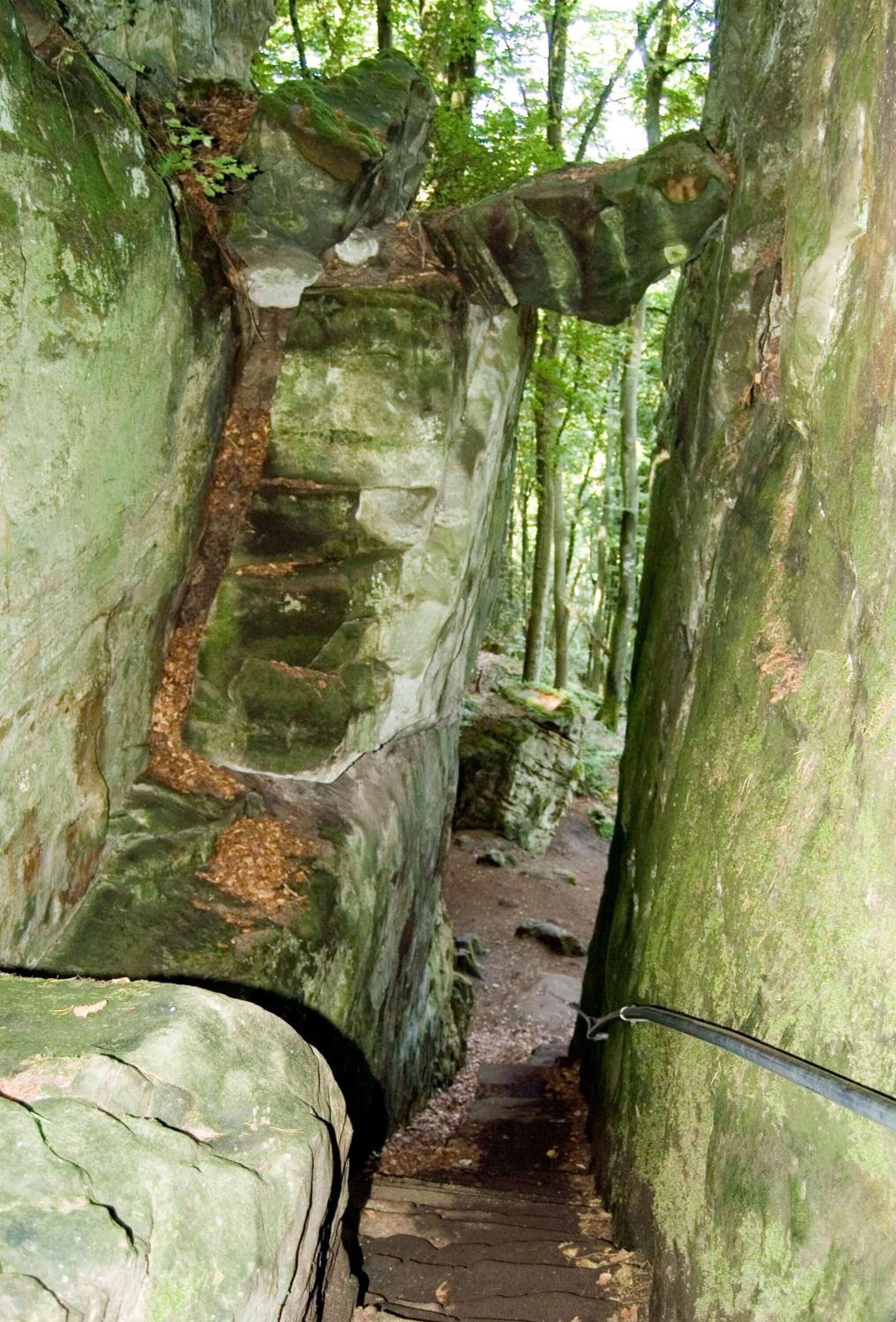 A narrow, rocky path flanked by large, moss-covered boulders in a forested area.