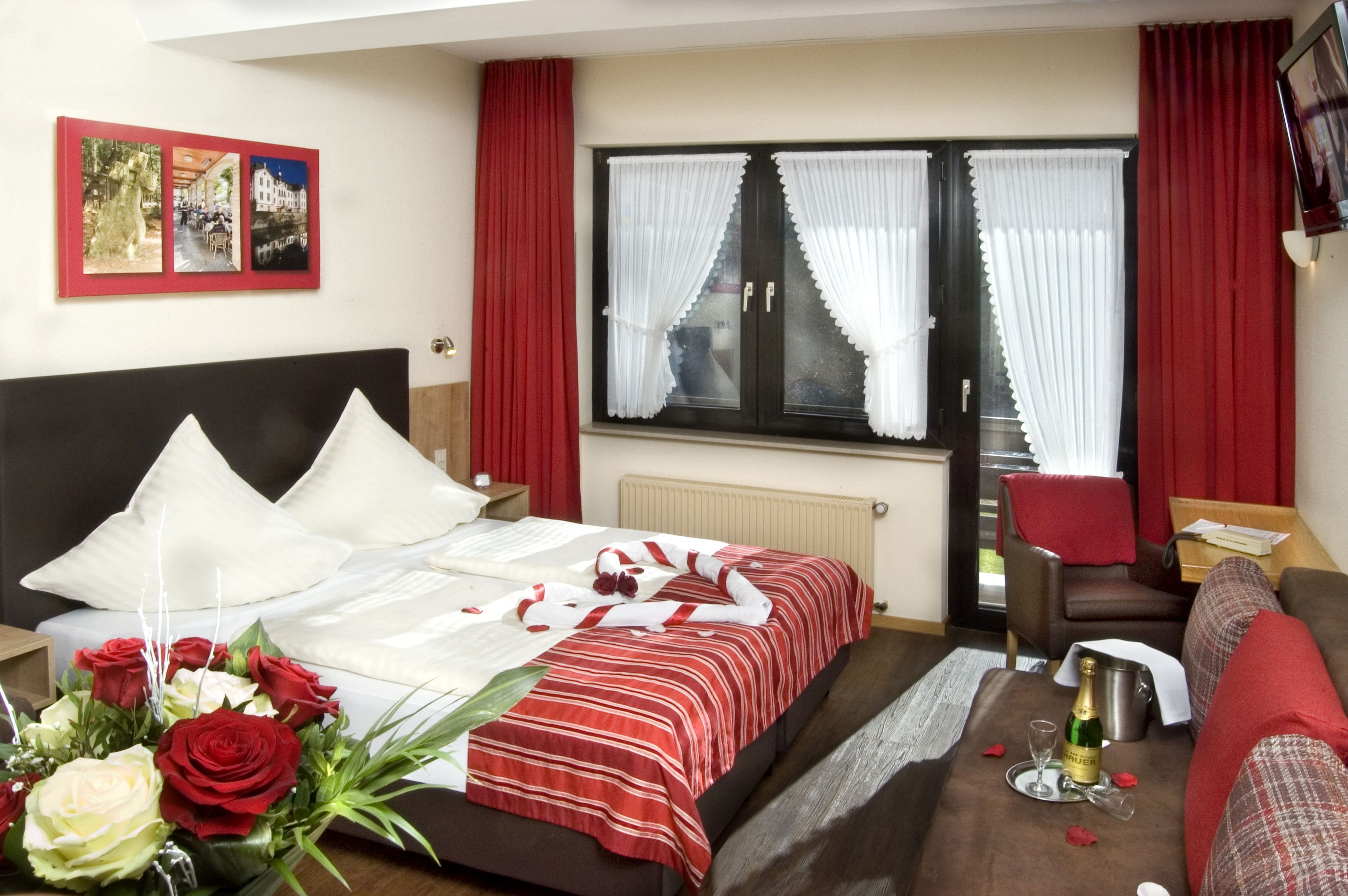 Ein Hotelzimmer mit einem Bett, einer rot-weiß gestreiften Tagesdecke, einem Strauß roter und weißer Rosen, einem roten Sessel, einem Tisch mit einer Champagnerflasche und einem roten Vorhang.